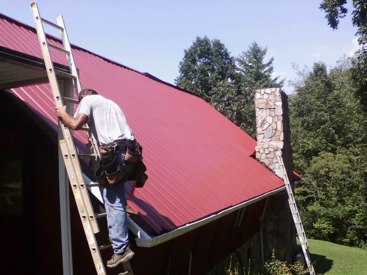 Roofer on a ladder beside a red metal roof and stone chimney — Soffit & Fascia Repair by Roofing Brothers in Battleboro